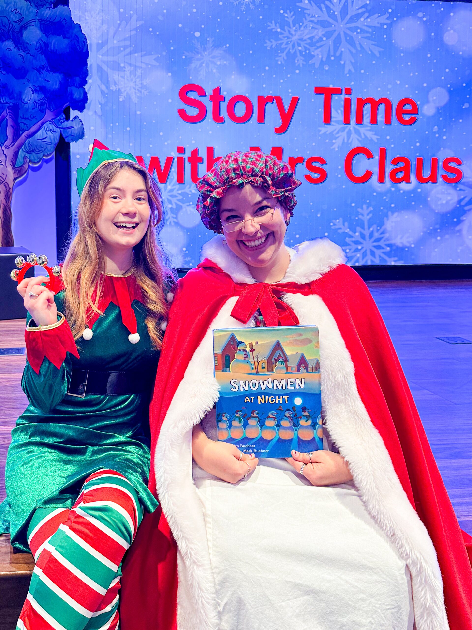 Mrs. Claus and elf sitting on the stage with bells and book in Story Time Theater